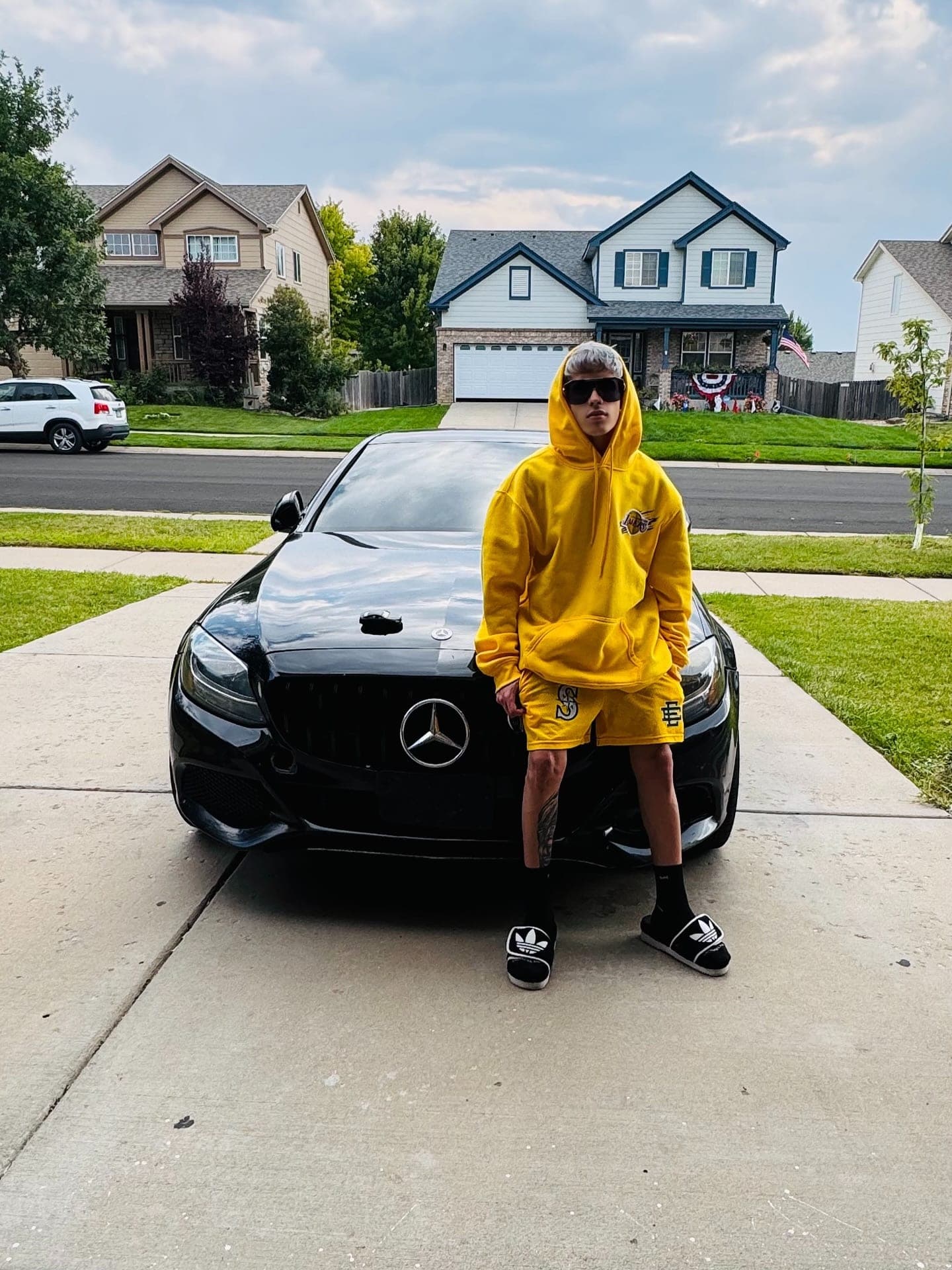 Milian con hoodie amarillo apoyado en un Mercedes negro en la entrada de una casa